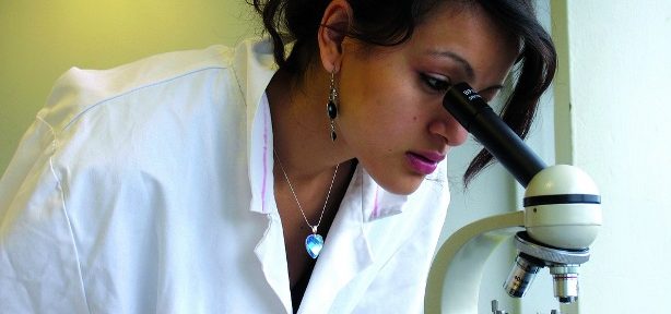 A young Asian female scientist wearing a white lab coat looks into a microscope