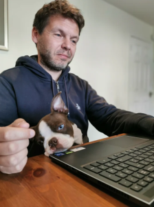 Fabien sitting in front of an open laptop looking at the screen with a dog sitting on his lap. 