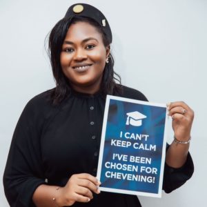 A woman smiles and holds up a sign that reads 'I can't keep calm, I've been chosen for Chevening!'