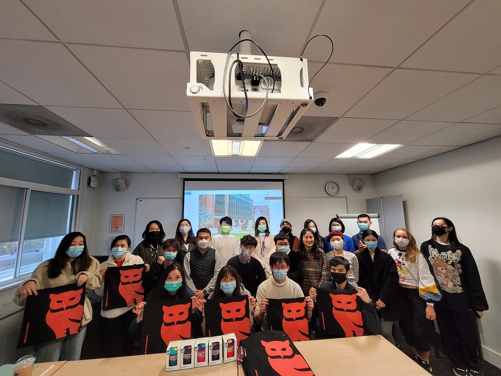 Students in  classroom with masks on, the people in the front row are holding up a branded bag with an orange owl on the front 