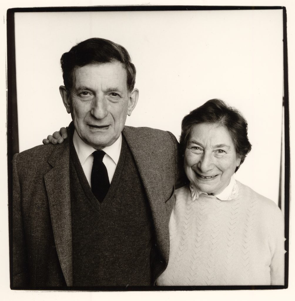 Black and white photo showing David and his wife. He wears his suit jacket, jumper, shirt and tie. They both smile at the camera.