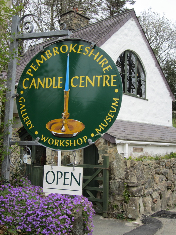 A circular dark green sign for the Pembrokeshire Candle Centre hangs in front a building with mauve flowers below.