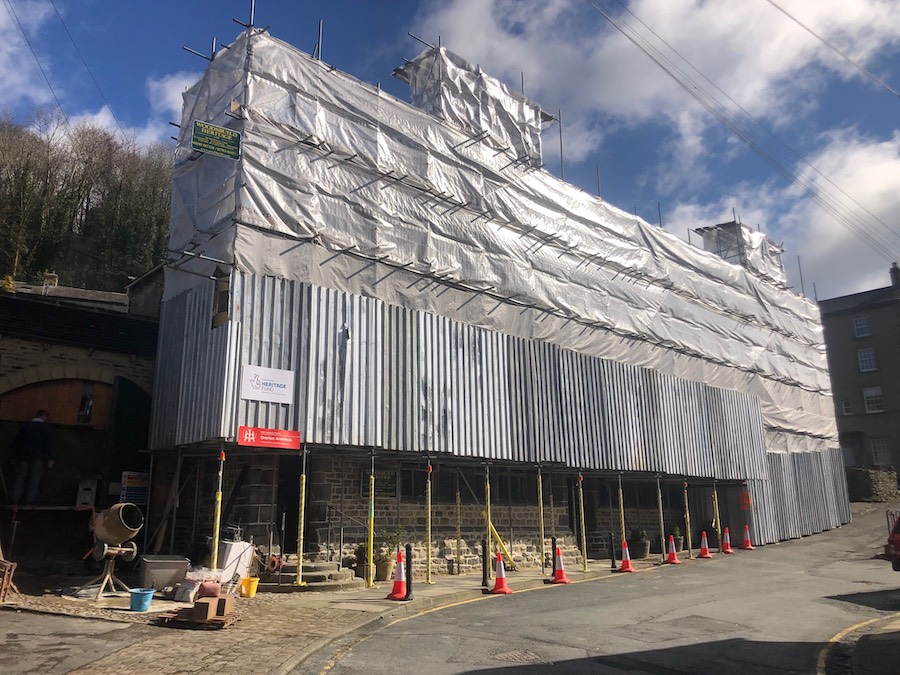 A large building covered in scaffolding, the uuper part of which is wrapped in plastic sheeting that reflects the sunlight. A sign for the Heritage Lottery Fund is affixed to the bottom left of the scaffold. A road runs past, leading off to the right.