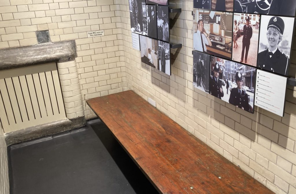 A cell inside the Bow Street Police Museum. Above a narrow dark wooden bench are displays of photographs and text.