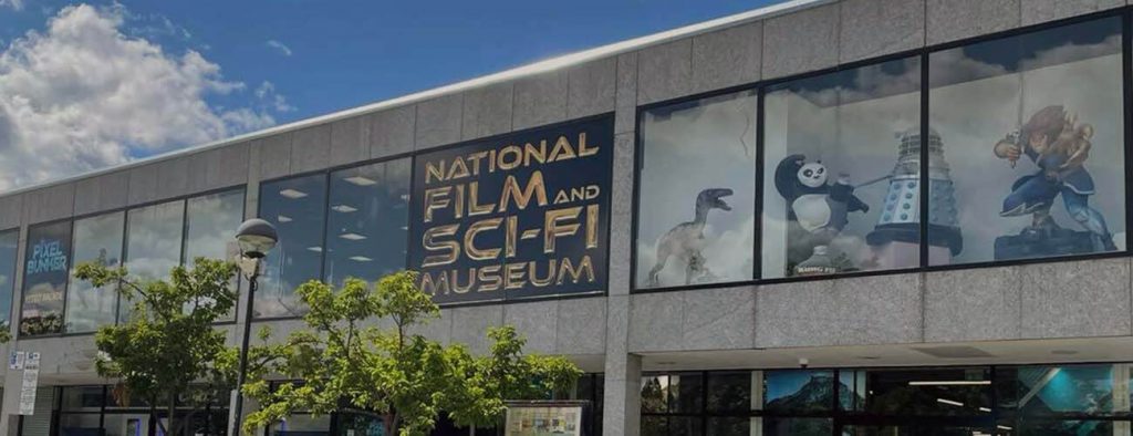 Exterior of the National Film and Science Fiction Museum in Milton Keynes. A two-storey building in stone or concrete, with large windows. Through an upstairs window can be seen figures including a dinosaur, Kung Fu Panda and a silver dalek.