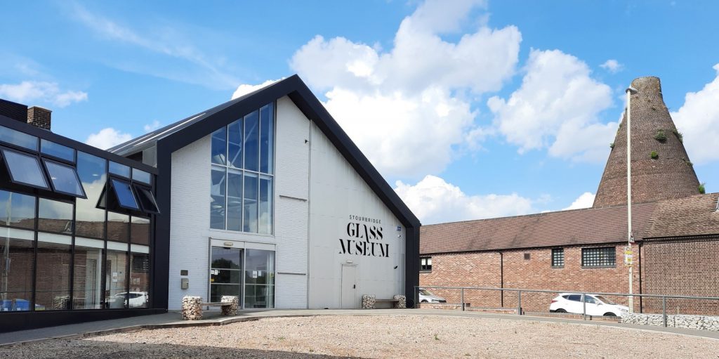 The front of Stourbridge Glass Museum, a modern building with white walls, glass frontage to the left. To the right an older brick building with a conical brick kiln rising above it.
