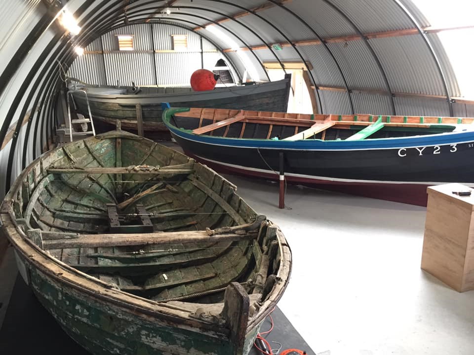 Interior of the boat shed at Grimsay Boat Haven. From front to back, a weathered dark green wooden dinghy, a much newer boat painted in dark blue and red, and a third boat with a rubber buoy resting on top.